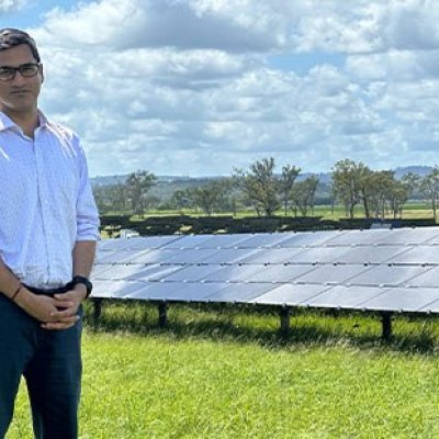  A man stands in a grassy field with rows of solar panels behind him. 